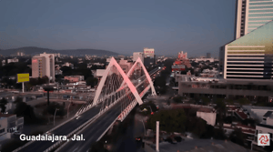 Vista aérea al atardecer del puente atirantado Matute Remus en Guadalajara, Jalisco. La estructura blanca iluminada en tonos rosados cruza una avenida con tráfico ligero, rodeada de edificios urbanos y cielo oscuro.